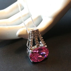 May include: A silver pendant necklace with a large, oval pink gemstone. The gemstone is set in a decorative, openwork frame with smaller, clear stones. The necklace has a fine chain. The pendant is a striking piece of jewellery.