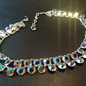 Op de afbeelding: Zilverkleurige ketting met twee strengen iriserende, druppelvormige kristallen. De kristallen reflecteren een spectrum aan kleuren, waaronder blauw, paars en geel. De ketting heeft een fijne ketting en een decoratieve sluiting.