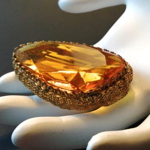Pode incluir: Anel com uma grande pedra preciosa ovalada de cor âmbar, com uma moldura de metal dourado texturizado. A pedra preciosa tem múltiplas facetas, refletindo a luz. Uma peça marcante para ocasiões especiais.