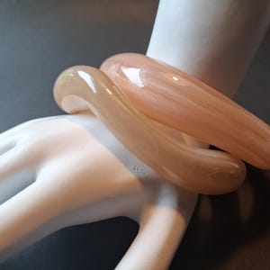 Op de afbeelding: Twee gladde, glanzende armbanden in tinten van bleek perzik en doorschijnend beige. De armbanden hebben een afgerond, dik profiel met een gepolijste afwerking die het licht reflecteert en een subtiele gemarmerde textuur in het materiaal toont.