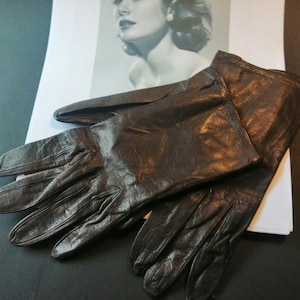 May include: A pair of dark brown leather gloves. The gloves are lying on a dark surface, with a black and white image in the background. The gloves have a classic design with detailed stitching.
