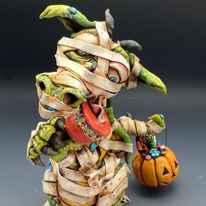Pode incluir: Uma estatueta de Halloween de uma criatura verde e bege envolta em ligaduras, segurando uma pequena cesta de abóbora laranja cheia de doces coloridos. A criatura tem uma língua vermelha e olhos azuis.