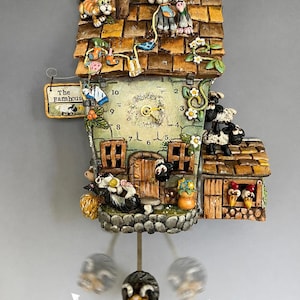 May include: A detailed farmhouse-style cuckoo clock. The clock has a rustic roof, a clock face with Roman numerals, and decorative animal figurines, including a cat, sheep, and cow. The pendulum has a cow face.