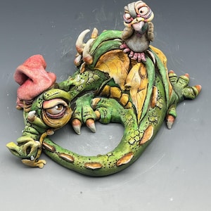 May include: A whimsical ceramic sculpture of a green dragon with yellow and orange accents, and a small bird perched on its back. The dragon has a pink hat and a detailed, expressive face. The sculpture is on a gray background.