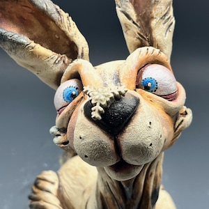 May include: A quirky ceramic rabbit sculpture with oversized features. The rabbit has large, expressive blue eyes, a black nose, and a snowflake-like detail. The sculpture is mainly beige with darker shading, giving it a textured appearance.