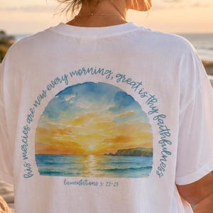 May include: A white short-sleeved t-shirt featuring a circular watercolour graphic on the back. The design depicts a sunset over the ocean with the text "his mercies are new every morning, great is thy faithfulness" and "Lamentations 3:22-23".