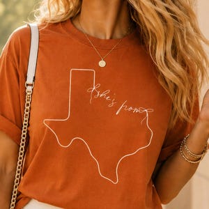 May include: A rust-colored short-sleeve t-shirt featuring a white line-art outline of the state of Texas with the cursive text "she's from" written inside the shape, paired with a white pleated skirt and a white shoulder bag with a gold chain.