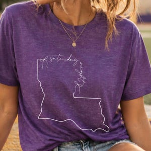 May include: A purple short-sleeved t-shirt featuring a white line-art outline of the state of Louisiana with the cursive text Saturday Night printed inside the state border, paired with light blue denim shorts and gold-toned jewelry.