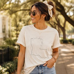 May include: A cream-colored short-sleeve t-shirt featuring a thin black outline of the state of Louisiana on the front. The graphic includes cursive text written vertically along the state border that reads Saturday in the South.