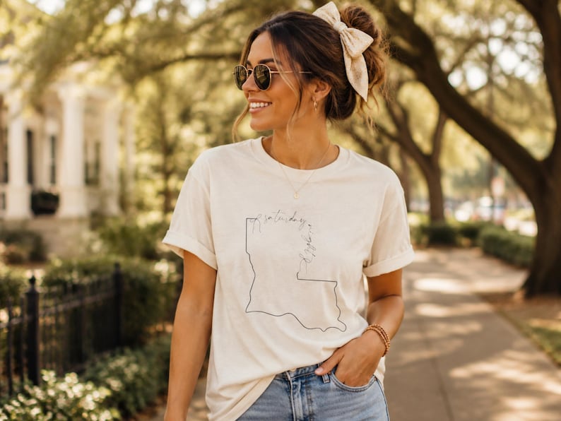 May include: A cream-colored short-sleeve t-shirt featuring a thin black outline of the state of Louisiana on the front. The graphic includes cursive text written vertically along the state border that reads Saturday in the South.