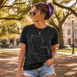 May include: A black short-sleeved t-shirt featuring a white line-art graphic of the state of Louisiana. The graphic includes the cursive text "saturday night" integrated into the outline of the state map, positioned centrally on the chest.