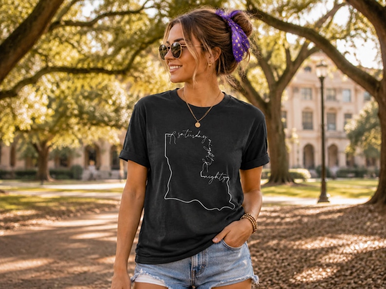 May include: A black short-sleeved t-shirt featuring a white line-art graphic of the state of Louisiana. The graphic includes the cursive text "saturday night" integrated into the outline of the state map, positioned centrally on the chest.