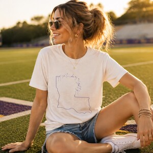 May include: A cream-colored short-sleeve t-shirt featuring a thin blue line drawing of the state of Louisiana. The graphic includes cursive text that reads Saturday in the South, positioned over the outline of the state map.