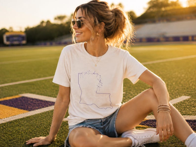 May include: A cream-colored short-sleeve t-shirt featuring a thin blue line drawing of the state of Louisiana. The graphic includes cursive text that reads Saturday in the South, positioned over the outline of the state map.