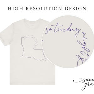 May include: A light beige short-sleeve t-shirt featuring a dark purple line art outline of the state of Louisiana. The text "saturday night" is written in a cursive script along the top edge of the state outline. A circular inset shows a magnified view.