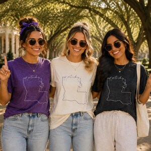 May include: Three short-sleeve crew neck t-shirts in purple, cream, and charcoal gray. Each shirt features a white line-art outline of the state of Louisiana with the cursive text "saturday night" integrated into the design on the front chest.