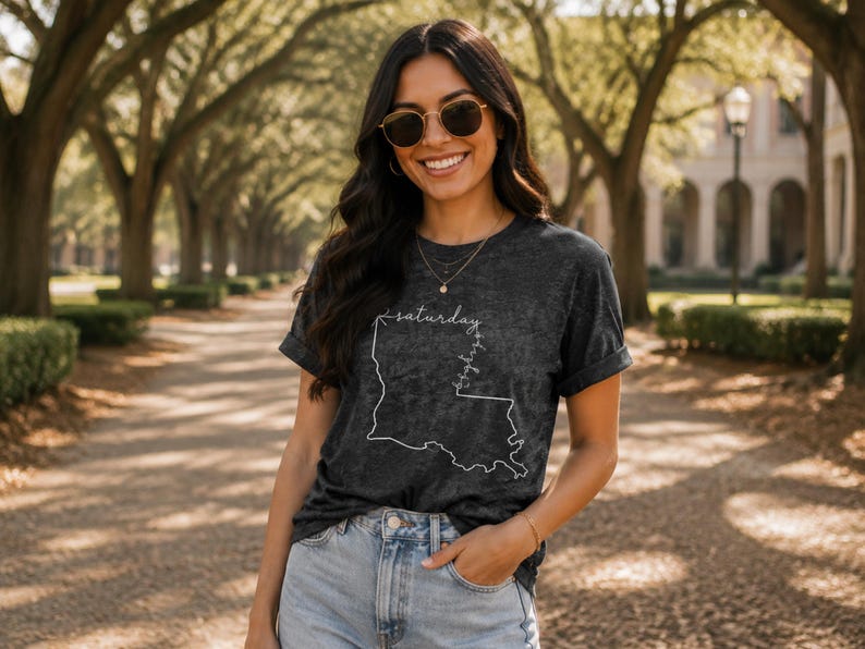 May include: A charcoal gray mineral wash t-shirt featuring a white line art outline of the state of Louisiana with the word saturday written in cursive script along the top border and the word bayou written vertically along the right side.