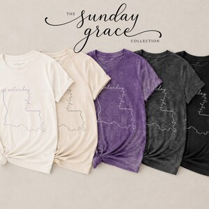 May include: Five short-sleeve t-shirts in cream, light beige, purple, dark gray, and black. Each shirt features a white line-art outline of the state of Louisiana with the word Saturday written horizontally and Sunday written vertically.