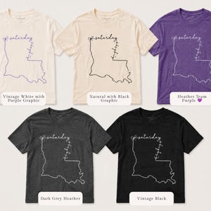 May include: Five short-sleeve crew neck t-shirts in vintage white, natural, heather team purple, dark grey heather, and vintage black. Each shirt features a line art graphic of the state of Louisiana with the word saturday and a bow.