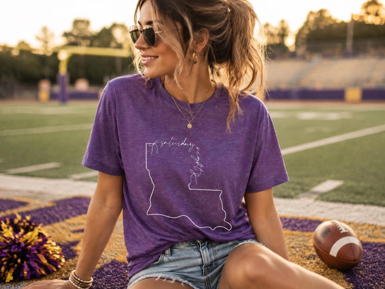 May include: A purple short-sleeved t-shirt featuring a white line-art outline of the state of Louisiana with the cursive text Saturday Night printed inside the state border, paired with light blue denim shorts and gold-toned jewelry.