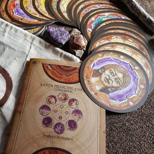 May include: A deck of circular oracle cards with colorful illustrations and a tan guidebook titled Earth Medicine Cards Guidebook Southern Hemisphere. The cards feature various artistic designs, including a portrait labeled Mother.