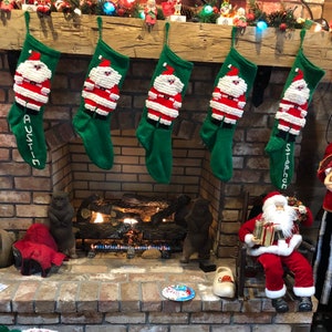 May include: Five red and green knitted stockings with Santa Claus designs hang on a brick fireplace mantel. The stockings are decorated with white trim and have the names "Austin" and "Stephen" written on them. A fireplace with a roaring fire is in the background.