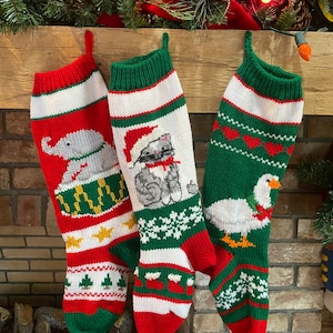 May include: Three knitted Christmas stockings hanging on a fireplace mantel. The stocking on the left features a red and green design with a white elephant playing a drum. The middle stocking is green and white with a gray cat wearing a Santa hat. The stocking on the right is green and white with a white goose wearing a red and green scarf.
