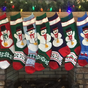 May include: Seven knitted Christmas stockings hang above a fireplace. Each stocking features a snowman design with a black top hat, red scarf, and broom. The stockings are red, green, blue, and navy, with white and green snowflake patterns.