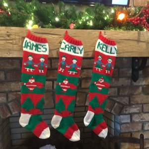 May include: Three red and green knitted Christmas stockings with white trim, hanging on a brick fireplace mantel. The stockings are personalized with the names "James", "Carly", and "Kelly".