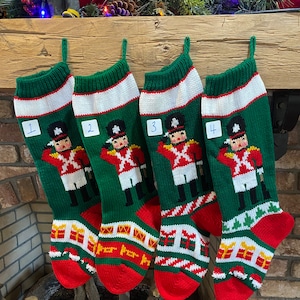 May include: Four green Christmas stockings with a white cuff and red toe are displayed. Each stocking features a knitted design of a toy soldier in a red coat and black hat. The stockings are numbered 1 through 4.