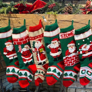 Peut inclure: Six bas de Noël tricotés, verts, rouges et blancs, suspendus à une cheminée en bois. Chaque bas présente un motif de Père Noël. Le nom "STONE" est tricoté sur l'un d'eux.