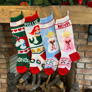 May include: Four knitted Christmas stockings hanging on a fireplace mantel. The stockings are decorated with different designs, including a unicorn, a mermaid, an angel, and a pink angel. The stockings are personalized with the names "Millie", "Nova", and two other names that are not visible in the image.