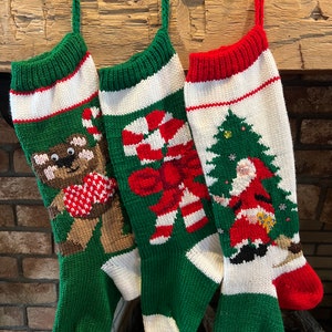 May include: Three knitted Christmas stockings with different designs. The stocking on the left has a brown teddy bear with a red heart, the middle stocking has a red and white candy cane, and the stocking on the right has a green Christmas tree with Santa Claus.