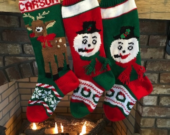 May include: Three knitted Christmas stockings hanging over a fireplace. The stocking on the left is green with a reindeer design and the name "CARSON" on it. The middle stocking is red with a snowman design. The stocking on the right is green with a snowman design.