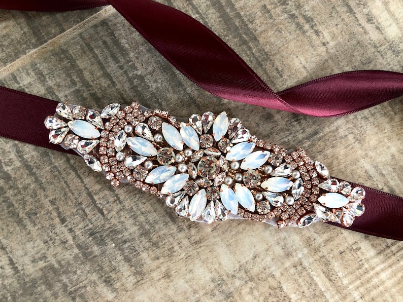 Puede incluir: Una cinta de sat&eacute;n burdeos con un adorno grande, ornamentado, de oro rosa y cristal. El adorno presenta cristales transparentes y blancos, as&iacute; como peque&ntilde;as perlas.