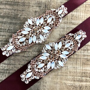 Puede incluir: Dos fajas de sat&eacute;n burdeos con adornos de strass de oro rosa y transparentes. Los adornos tienen un patr&oacute;n floral.