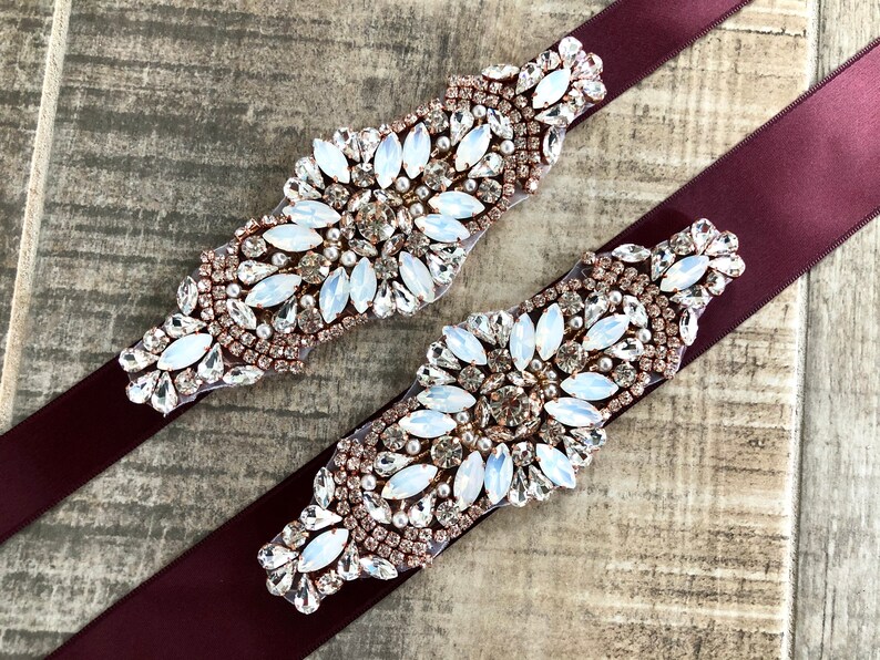 Puede incluir: Dos fajas de sat&eacute;n burdeos con adornos de strass de oro rosa y transparentes. Los adornos tienen un patr&oacute;n floral.