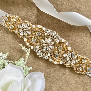 Puede incluir: Una cinta de satén blanco con una hebilla de cinturón adornada con oro y cristales. La hebilla presenta un diseño floral con diamantes de imitación transparentes y detalles de perlas.