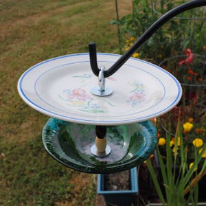 May include: A bird feeder made from two plates, one white with blue trim and floral designs, and the other green glass. The feeder is suspended from a black metal hook.