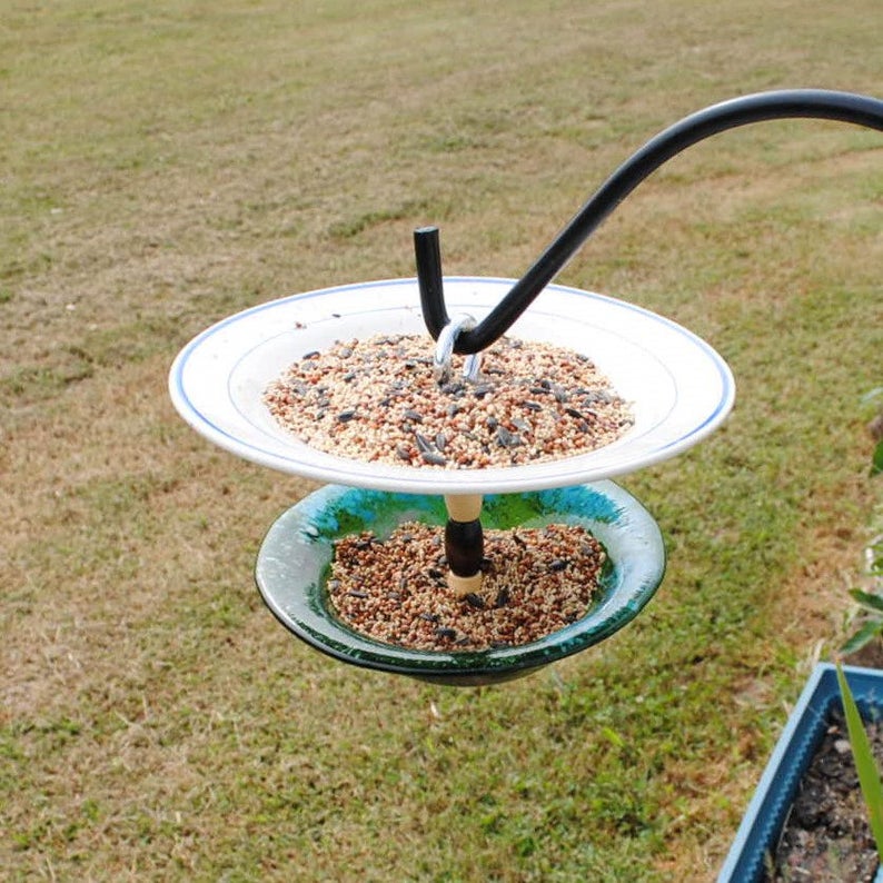 May include: A bird feeder made from two plates, one white and one blue, with a black metal hook. The feeder is filled with birdseed and is hanging from a black metal hook.