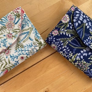 May include: Two fabric pouches with floral patterns. One pouch is white with pink and blue flowers and green leaves. The other pouch is navy blue with purple and pink flowers and green leaves.