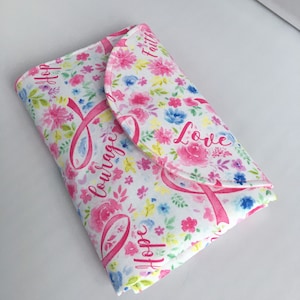 May include: A white fabric clutch with a floral pattern and pink ribbons. The words "Faith", "Love", "Hope", and "Courage" are printed on the clutch.