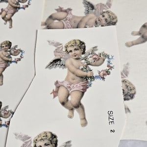 Könnte beinhalten: Eine Vintage-Aufkleberfolie mit mehreren Bildern von Cherubim, die Blumenkränze halten. Die Cherubim sind in einem zarten, verspielten Stil mit Pastellfarben dargestellt. Die Aufkleberfolie ist mit "SIZE 2" beschriftet.