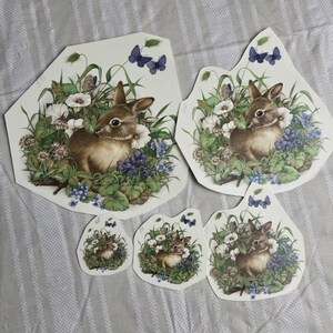 Puede incluir: Cinco recortes de papel de un conejo marrón rodeado de flores azules y moradas. El conejo está sentado en un campo de hierba verde. Los recortes son de diferentes tamaños y son perfectos para manualidades o scrapbooking.
