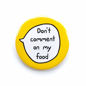 May include: A round, yellow button with the text "Don't comment on my food" inside a white speech bubble outlined in black. The button is a fun, quirky accessory.