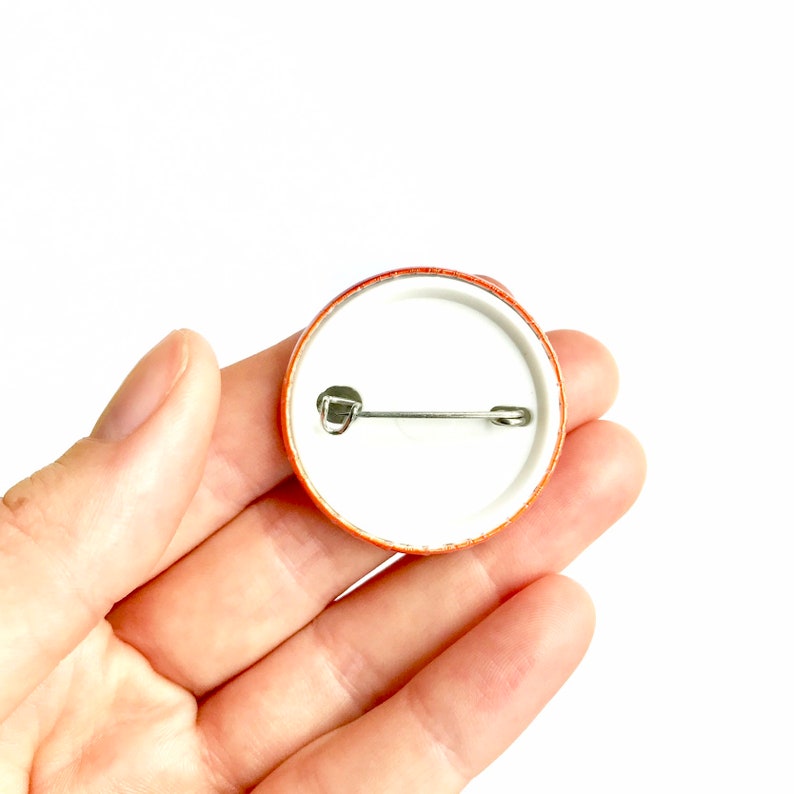 May include: The back of a round button with a safety pin attached. The button is orange and white.