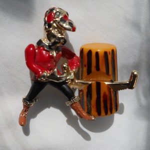 May include: A vintage brooch featuring a jester figure in red, black, and gold enamel, leaning against a barrel. The jester has a detailed face and is wearing a red jacket and black pants. The barrel is orange with brown stripes.