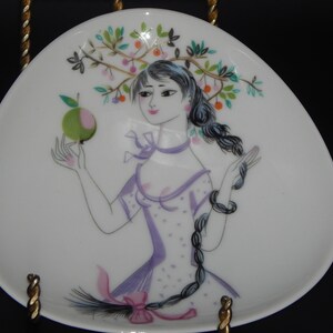 May include: White, teardrop-shaped decorative plate featuring a stylized illustration of a woman holding an apple. The woman has long black braids and wears a purple dress. The plate is decorated with a tree branch with fruit and leaves.