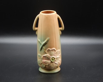 May include: A peach-colored ceramic vase with two handles and a floral design. The vase has a green and peach color gradient, with a flower and leaf design on the front. The vase is approximately 8 inches tall.