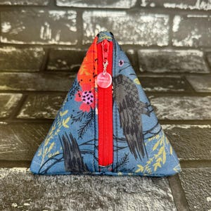 May include: A triangular zippered pouch with a blue background and a floral and bird print. The zipper is red and has a silver charm with the text "Made in the USA".